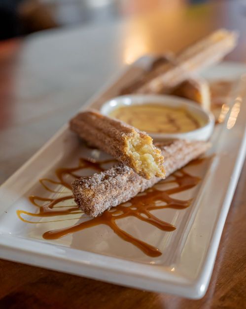 A plate of churros with a bite taken out, served with a creamy dipping sauce and drizzled with caramel.