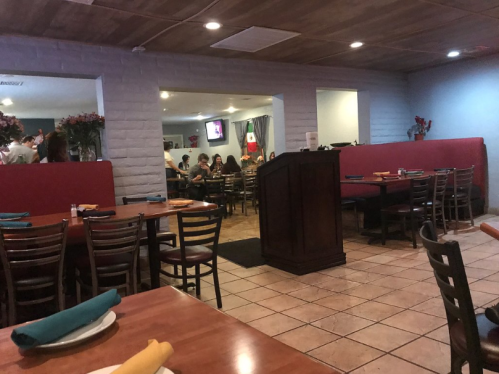 Interior of a restaurant with wooden tables, chairs, and a podium, featuring diners in the background and floral decorations.