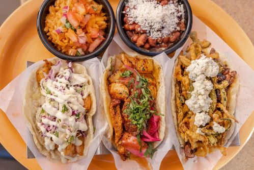 A colorful platter featuring three tacos with various toppings, accompanied by rice, pico de gallo, and beans.