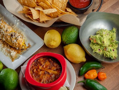 A vibrant spread of Mexican dishes: tortilla chips, guacamole, elote, and a bowl of soup, surrounded by fresh limes and peppers.