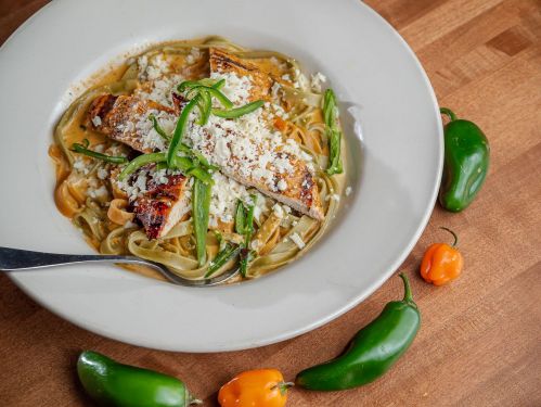 A plate of pasta topped with grilled chicken, cheese, and green peppers, surrounded by fresh chili peppers.
