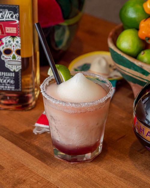A frosty cocktail with a salted rim, topped with foam and a lime wedge, surrounded by colorful fruits and bottles.