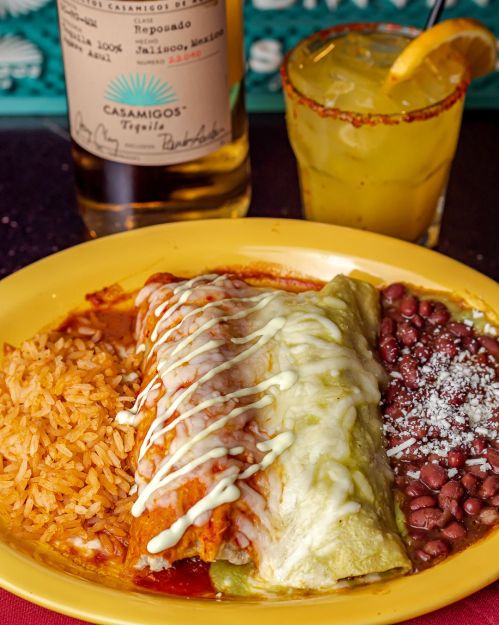 A plate of enchiladas with rice and beans, accompanied by a cocktail and a bottle of tequila in the background.