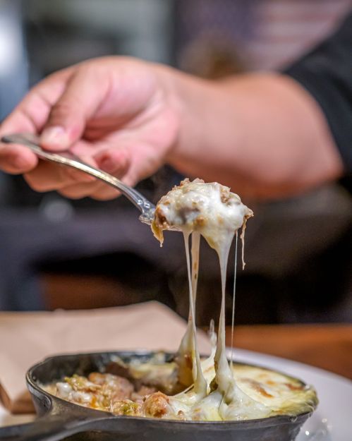 A hand holding a spoonful of cheesy, melted dish from a small skillet, with strings of cheese stretching.