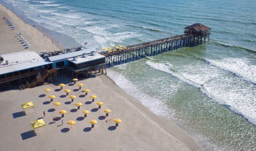 Aerial view of a beach with umbrellas, a pier, and gentle waves lapping at the shore.
