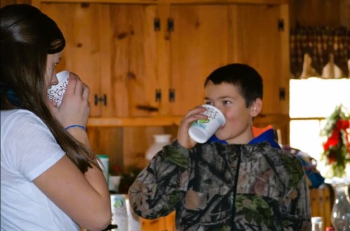 A boy and girl drink from cups in a cozy kitchen with wooden cabinets and festive decorations.