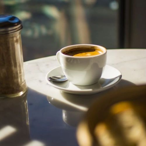 A cup of coffee on a saucer sits on a table, with a sugar jar nearby and sunlight casting soft shadows.
