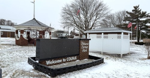 A brick monument sign welcoming visitors to Colfax and Mineral Springs Park stands in the snow, flanked by a historic gazebo and a white park building on a winter day.