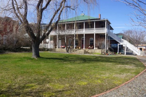 A two-story house with a green roof, white railings, and a large lawn, surrounded by trees and a gravel path.