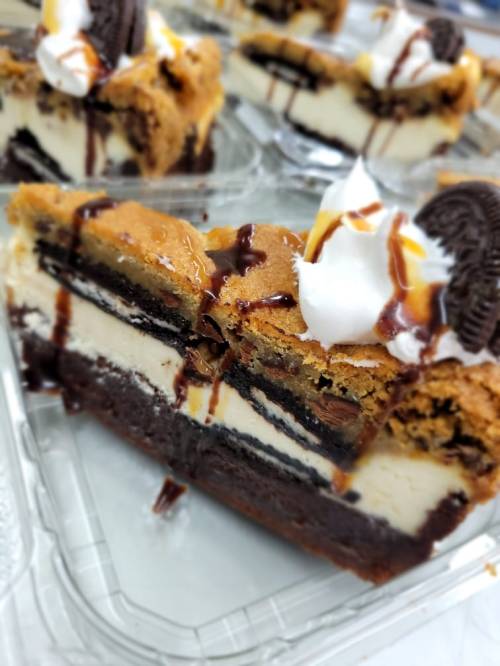 A slice of layered dessert featuring brownie, cheesecake, and cookie dough, topped with whipped cream and chocolate drizzle.