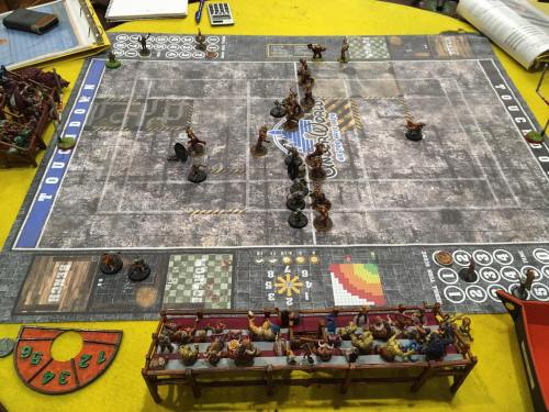 A tabletop game setup with miniatures on a textured game board, featuring a scoreboard and player stands.