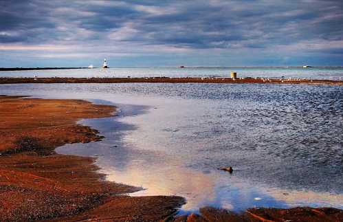 A serene beach scene with a lighthouse in the distance, reflecting clouds and water on the sandy shore.