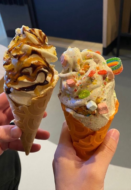Two ice cream cones held in hands: one with chocolate sauce and whipped cream, the other with colorful sprinkles and candy.