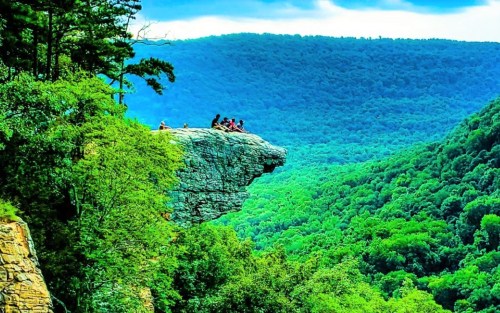 A rocky outcrop overlooks a lush green valley, with people sitting at the edge enjoying the scenic view.