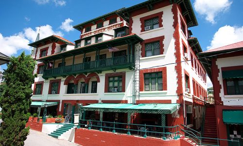 Historic hotel with red and white exterior, green balconies, and American flags, set against a blue sky.