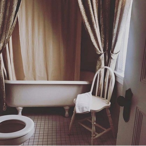 A vintage bathroom featuring a clawfoot tub, a wooden chair, and a toilet, with elegant curtains and natural light.