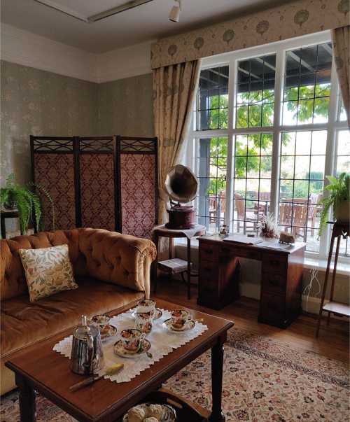 Cozy vintage living room with a sofa, tea set, wooden desk, and large windows overlooking greenery.