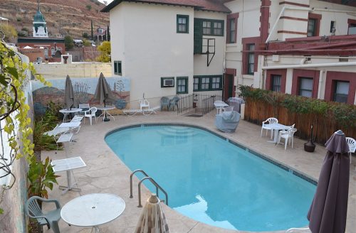 A view of a pool surrounded by lounge chairs and tables, with a building and greenery in the background.