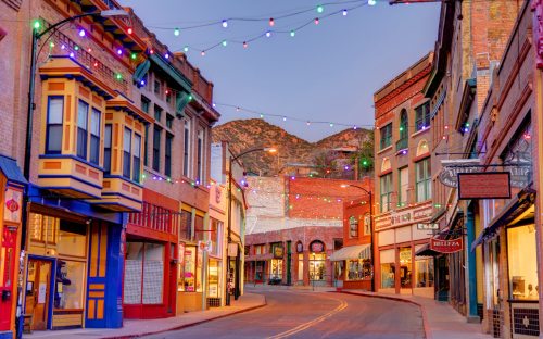 Colorful buildings line a quiet street adorned with festive lights, set against a mountain backdrop at dusk.
