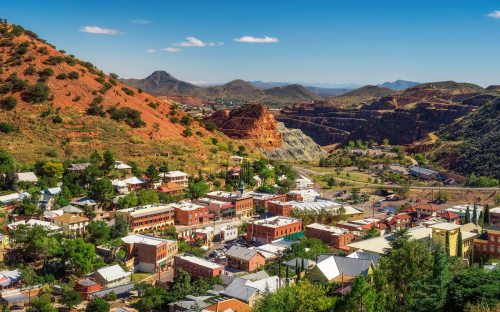 A scenic view of a small town nestled in mountains, with red rock formations and clear blue skies in the background.
