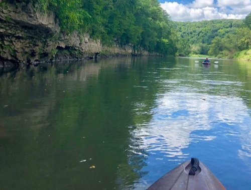 A serene river scene with lush green cliffs and a kayaker in the distance under a partly cloudy sky.