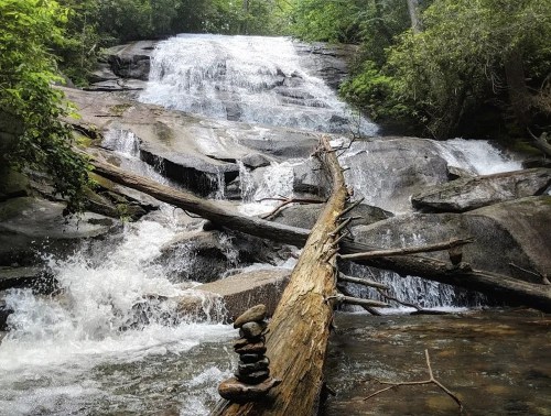 A serene waterfall cascades over rocks, with a log and stacked stones in the foreground, surrounded by lush greenery.