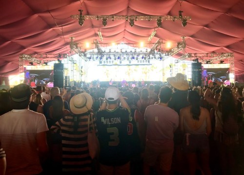 A crowd of festival-goers watches a performance on stage under a large, pink tent.