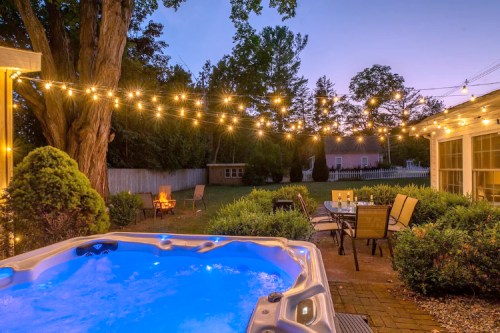 A serene backyard at dusk featuring a hot tub, string lights, and a cozy seating area surrounded by greenery.