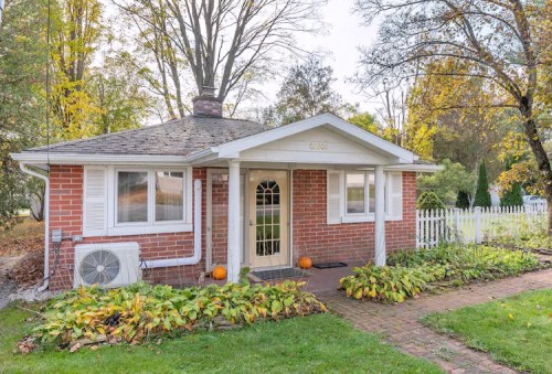 A cozy brick house with a white porch, surrounded by greenery and autumn leaves, featuring pumpkins by the entrance.