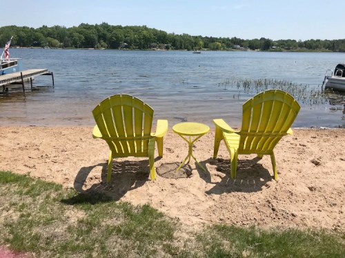 Two yellow Adirondack chairs and a small table on a sandy beach by a calm lake, surrounded by greenery.