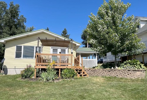 A yellow house with a wooden deck, surrounded by greenery and a tree, under a clear blue sky.