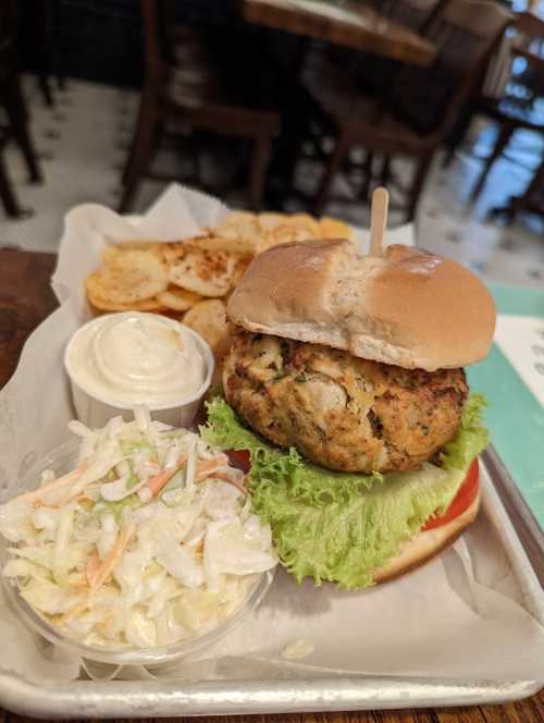 A plate featuring a crab cake sandwich on a bun, lettuce, a side of coleslaw, and crispy potato chips with dipping sauce.