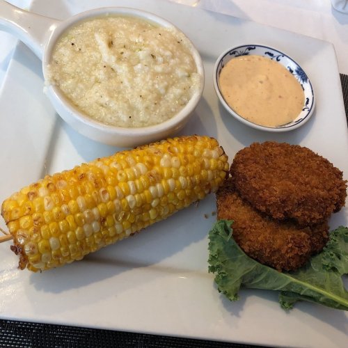 A plate with creamy grits, fried corn, two golden fritters, and a small bowl of dipping sauce.