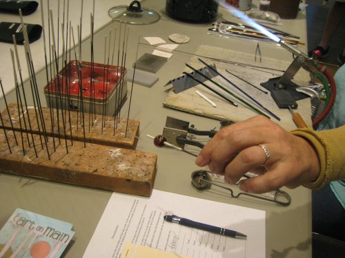 A hand reaches for tools on a workbench, surrounded by various metal rods, a pen, and papers.