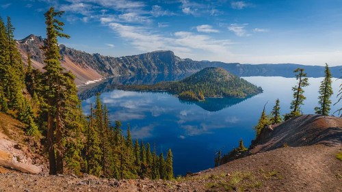A serene view of Crater Lake, surrounded by lush trees and mountains, reflecting a clear blue sky.