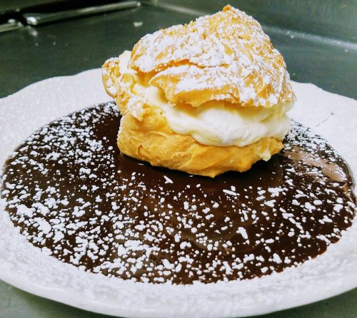 A cream puff topped with powdered sugar sits on a plate of rich chocolate sauce, dusted with more powdered sugar.
