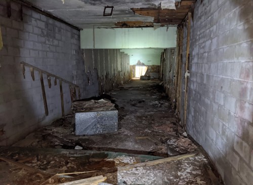 A dimly lit, abandoned hallway with debris and peeling walls, leading to a bright exit in the distance.