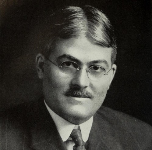 Black and white portrait of a man with glasses, a mustache, and slicked-back hair, wearing a suit and tie.