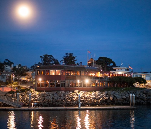 A waterfront restaurant illuminated at night, with a full moon shining above and reflections on the water.