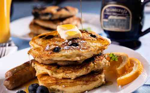 Stack of fluffy pancakes topped with butter and syrup, served with blueberries, sausage, and a slice of orange.