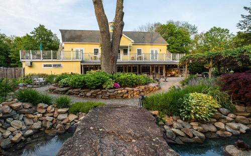 A yellow house with a deck surrounded by lush gardens and a stone pathway leading to a pond.