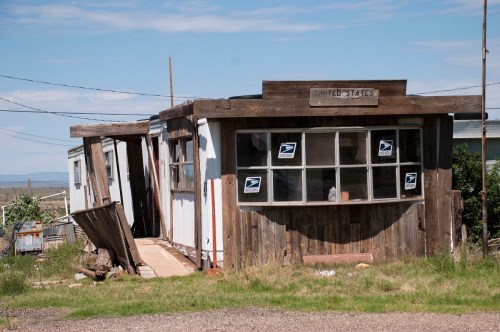 Abandoned post office building with wooden exterior and USPS signs, surrounded by grass and a clear blue sky.