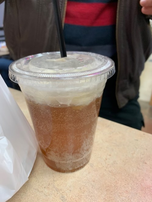 A clear plastic cup filled with a fizzy brown drink and ice, placed on a table next to a white bag.