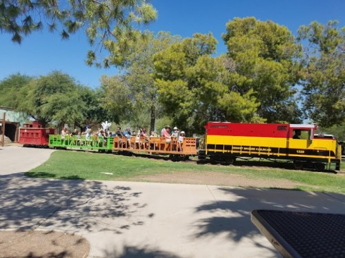 A colorful train with passengers rides through a park surrounded by trees on a sunny day.