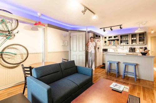 A cozy apartment interior featuring a sofa, dining area, kitchen, and a person standing by the door.