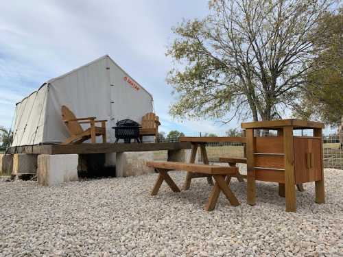 A glamping tent on a wooden platform, with picnic tables and a grill, surrounded by gravel and trees.