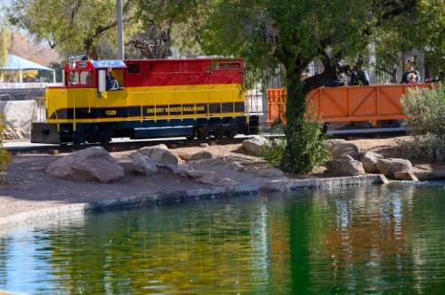 A colorful miniature train near a pond, surrounded by rocks and trees in a park setting.