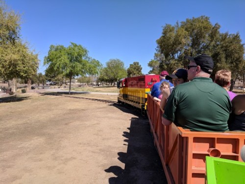 A colorful train with passengers rides along a track in a park, surrounded by trees and clear blue skies.