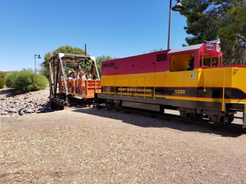 A colorful train with passengers on an open car, stopped on a gravel path surrounded by greenery.