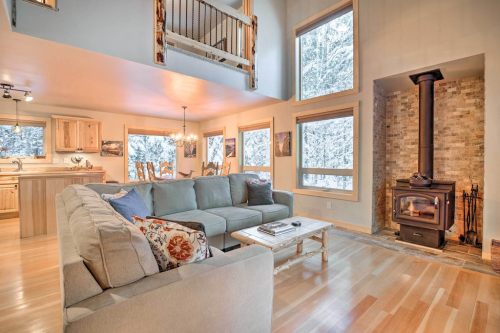 Cozy living room with a gray sofa, wooden accents, and a wood stove, featuring large windows with a snowy view outside.